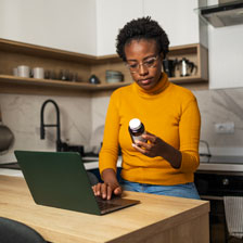 A woman sits at her kitchen counter reading a pill bottle while typing on her laptop