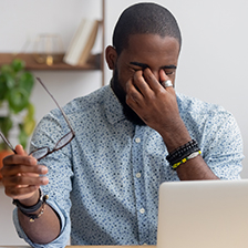 Man sits at a desk in front of a computer while rubbing his eyes