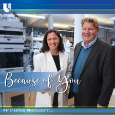 Two scientists, a woman in a white lab coat and a man in a sport coat, stand in a lab and smile at the camera. 