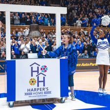 Harper Harrell, a 10-year-old Duke Children's cancer patient, rings a special bell to signal the end of her treatment. She is in Cameron Indoor Stadium at a Duke Men's Basketball game and is surrounded by fans and cheerleaders. 