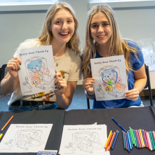 Two women holding coloring sheets in front a table with coloring materials 
