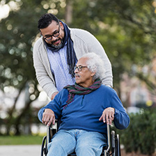 Man pushes another man in a wheelchair in an outdoor setting