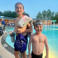 Two 9-year-old boys in swim gear stand in front of a pool and smile. 