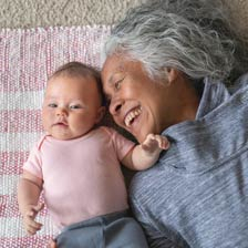 A baby in pink and an older lady dressed in blue lie on the floor and smile