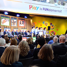 A choir sings while a panel of speakers and a crowd look on in front of a sign that reads Play is Fundamental