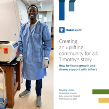 Timothy smiles in a blue lab coat next to a machine. 
