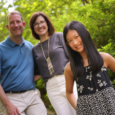 Violet Ake, a 13yo girl of Asian descent, stands in front of her parents (both white and middle-aged) and smiles. Violet has long dark hair and wears a black and white floral dress. She and her parents are in an outdoor garden setting, lots of greenery. 