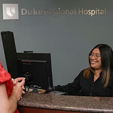 A woman sits at a reception desk smiling, under the Duke Regional Hospital logo